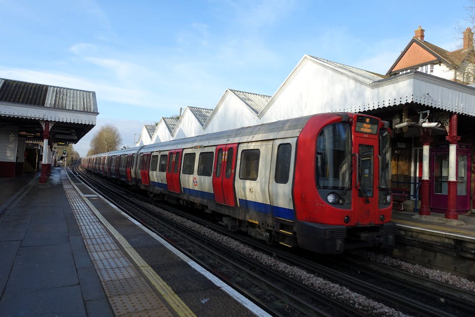 A man in a grey t-shirt and high-visibility orange vest stands on a metro station platform, facing away from the camera and looking towards an approaching yellow and black train. The platform surface
