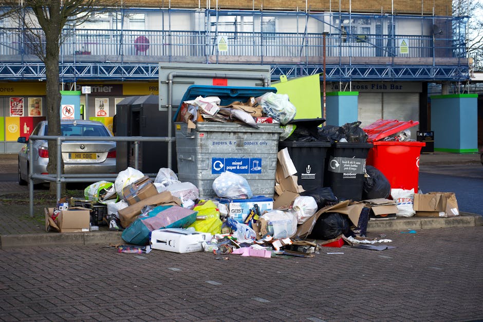 A collection of overflowing rubbish bins and scattered waste on a paved sidewalk in front of a retail building with a blue scaffolding structure. The waste includes cardboard boxes, plastic bags fille
