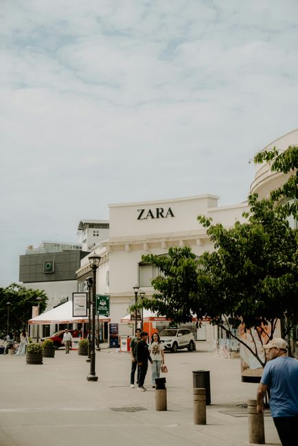 A collection of overflowing rubbish bins and scattered waste on a paved sidewalk in front of a retail building with a blue scaffolding structure. The waste includes cardboard boxes, plastic bags fille